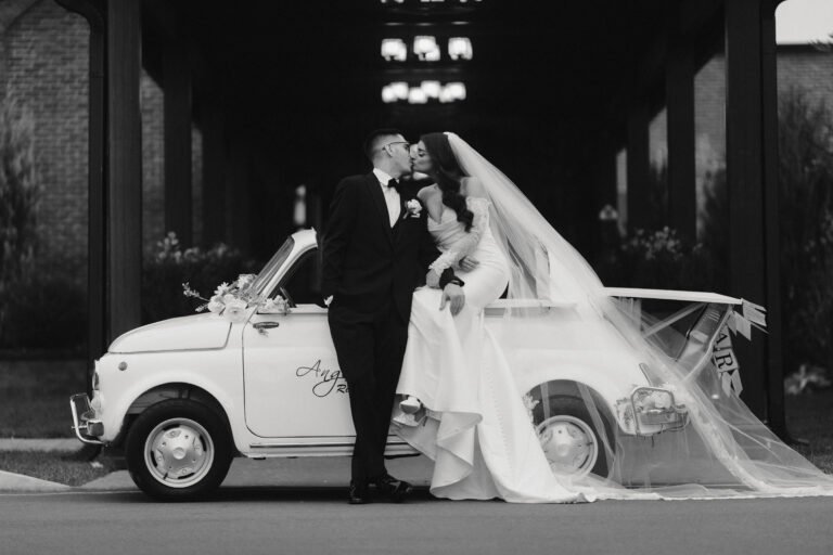 Black-and-white photo of a bride and groom kissing beside a vintage convertible getaway car