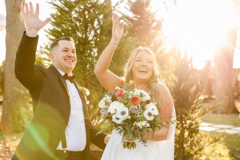 Bride and groom cheer with arms raised in golden sunlight on the grounds