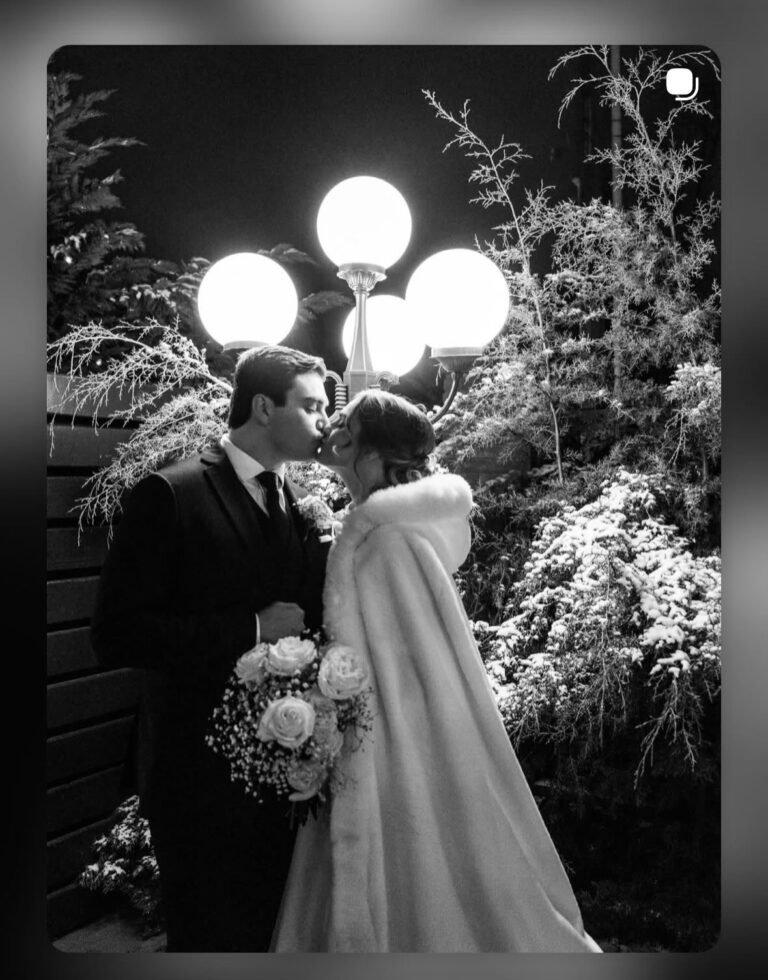 Black-and-white photo of a bride and groom kissing beneath a vintage globe lamppost at night