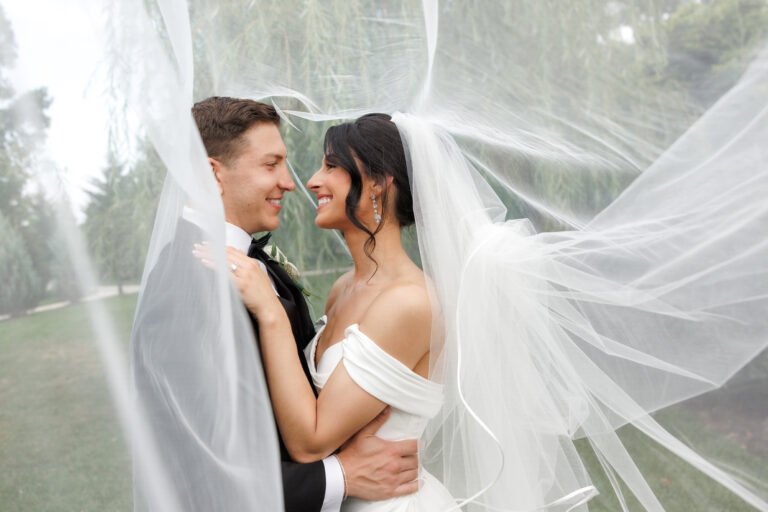 Groom and bride smile at each other as her veil billows around them outdoors