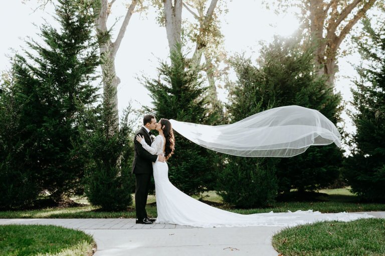 Bride's cathedral veil catches the wind as she kisses the groom on a garden path