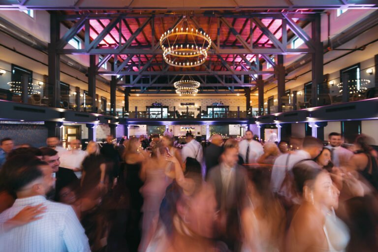 Guests dancing beneath a ring chandelier and exposed beams lit in purple and blue