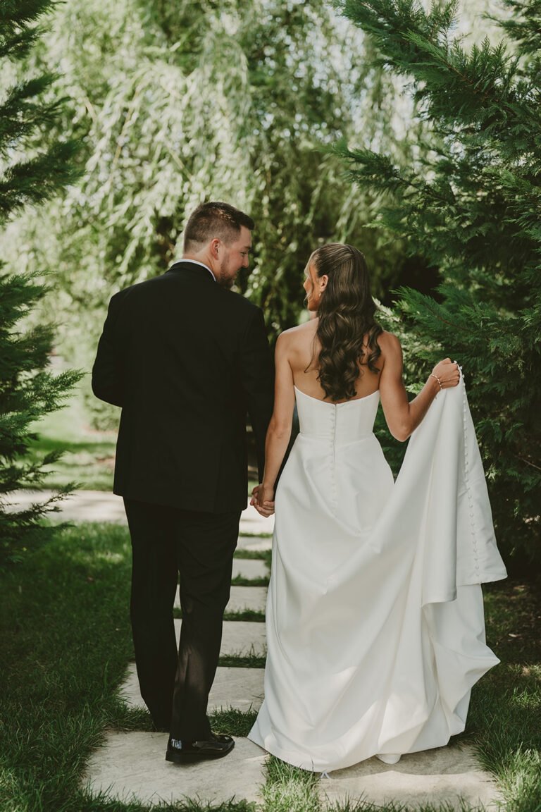 Bride and groom walk hand in hand along a stone path beneath willow trees