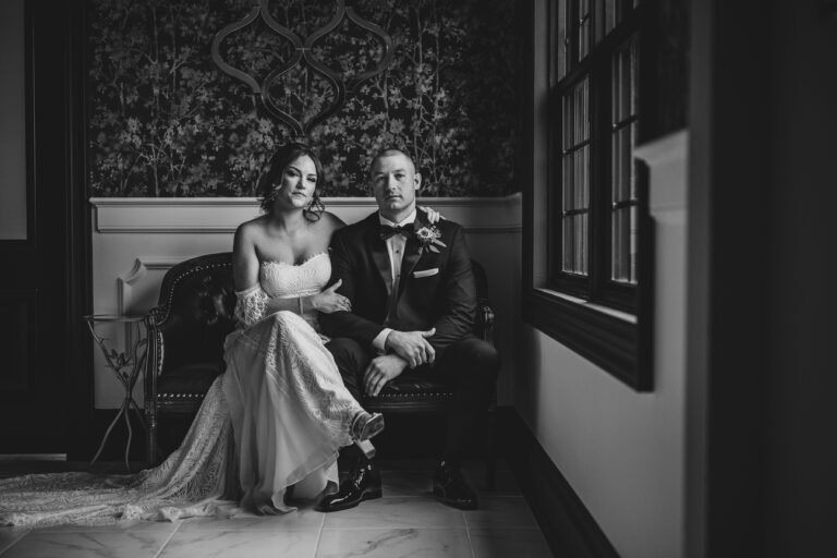 Black-and-white portrait of a bride and groom seated on a tufted bench in a wallpapered lounge