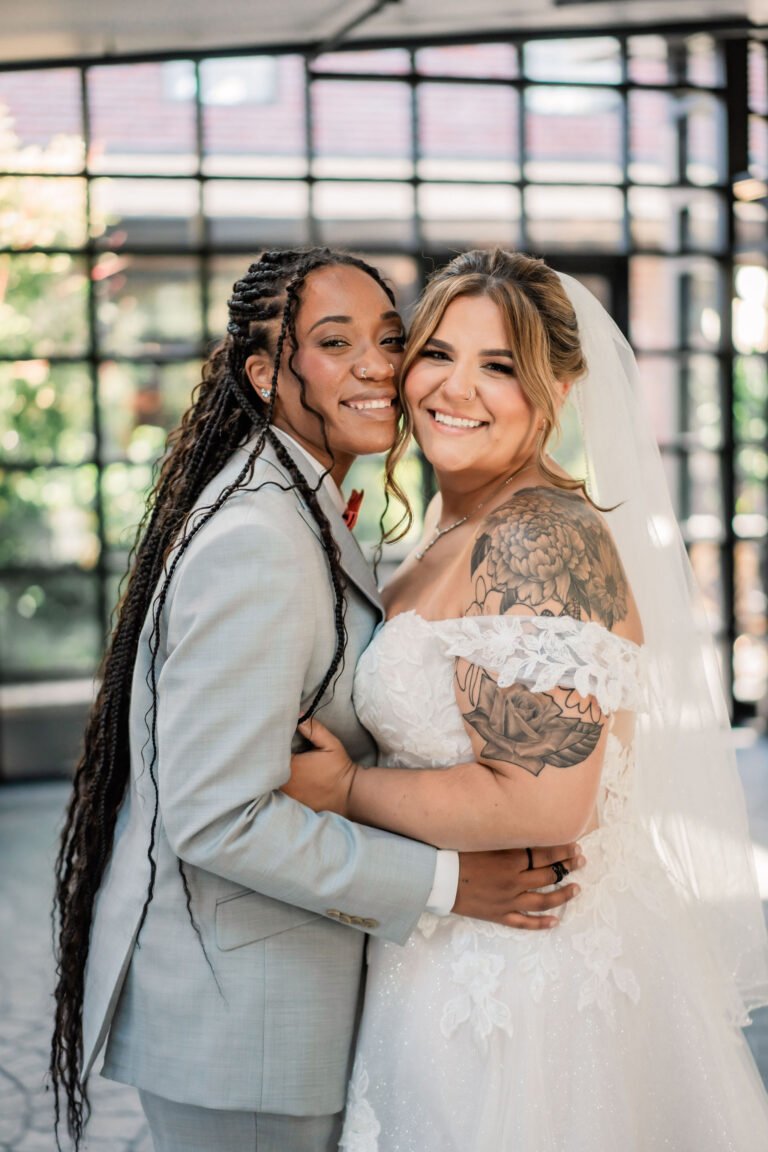 Two brides embrace and smile inside the glass-walled atrium at Park Loft