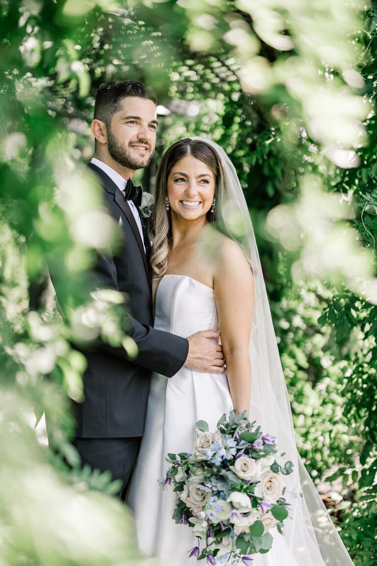 Bride and groom pose among lush greenery, the bride holding a white and blue bouquet