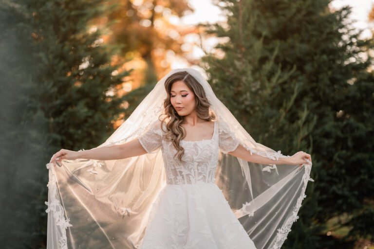 Bride in a lace gown spreads her cathedral veil at golden hour among the evergreens