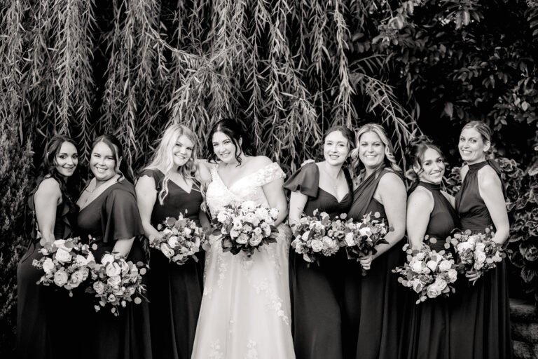 Black-and-white photo of the bride with bridesmaids holding bouquets beneath a weeping willow