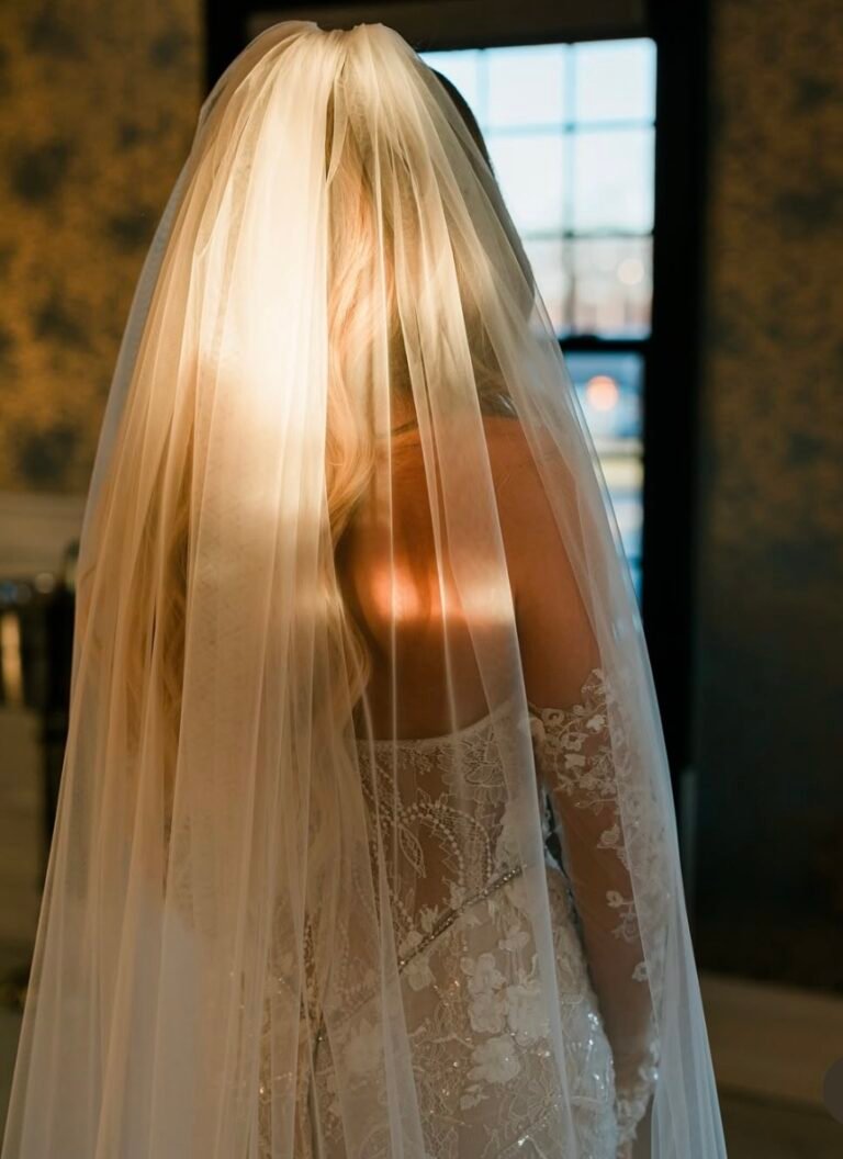 Sunlight glows through the bride's veil as she stands by a window
