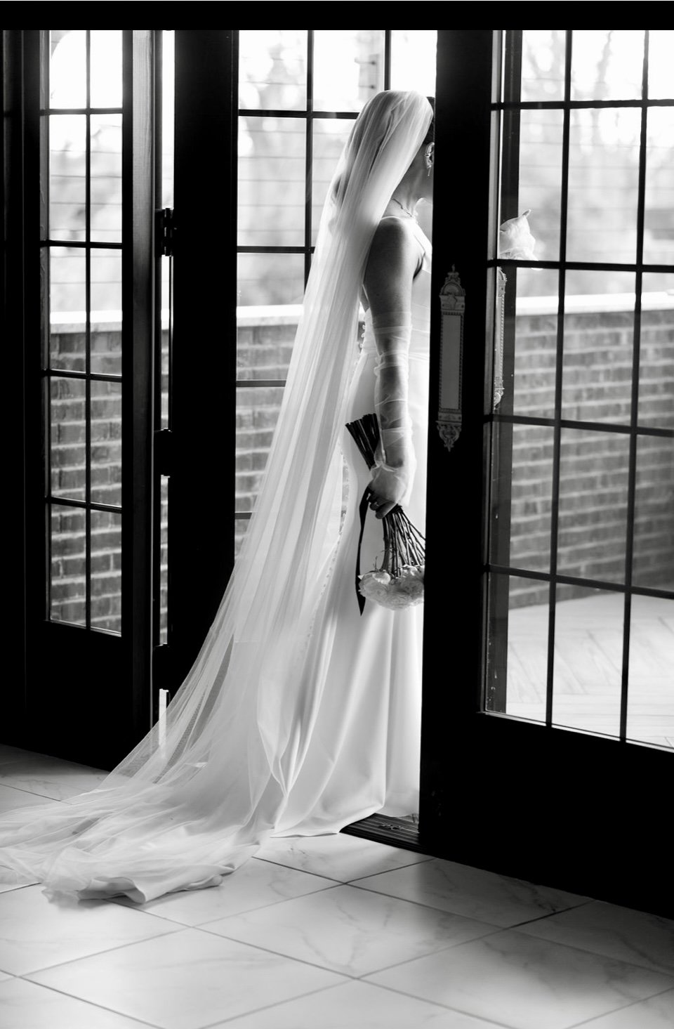 Black-and-white photo of a bride in a long veil standing at the French doors