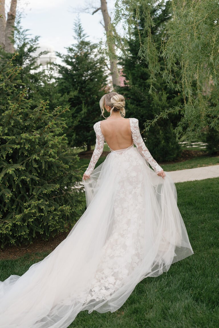 Bride in a long-sleeve lace gown with an open back walks across the lawn toward the evergreens