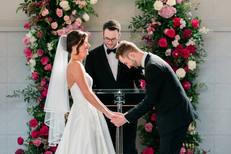 Couple holds hands at the altar beneath a pink and red rose floral arch during their Park Loft ceremony