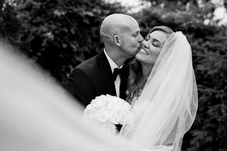 Black-and-white photo of a groom kissing the bride's cheek as she holds a white rose bouquet