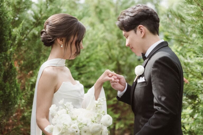 Groom holds the bride's hand among greenery as she carries a white bouquet