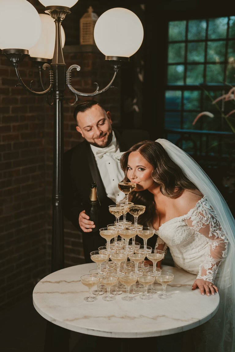 Bride sips champagne from a champagne tower beside the groom and a vintage globe lamp