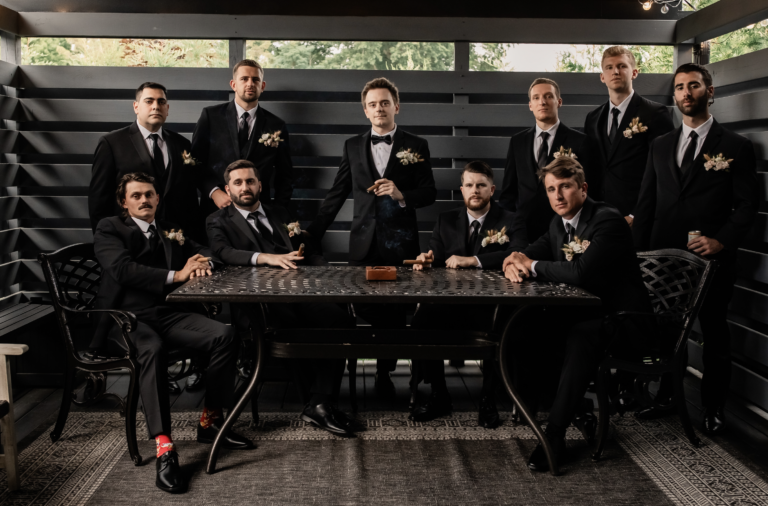 Groom and groomsmen in black tuxedos gathered around a table on the covered patio