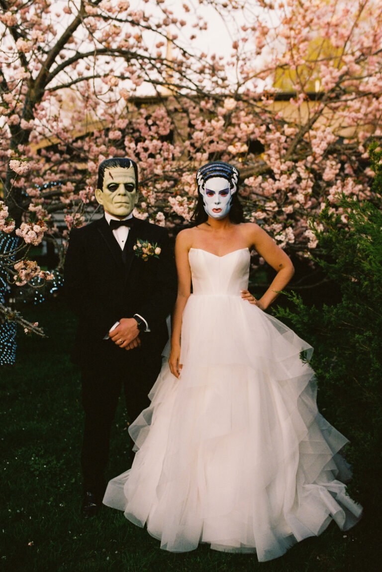 Playful portrait of a bride and groom wearing costume masks beneath cherry blossoms