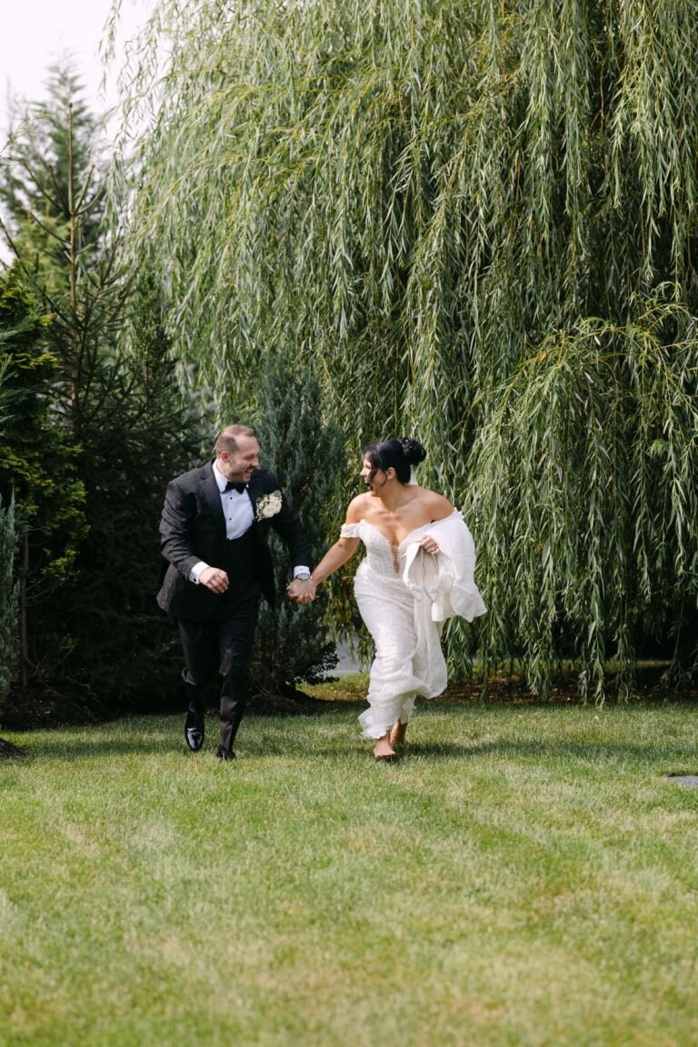 Bride and groom run hand in hand across the lawn beneath willow trees