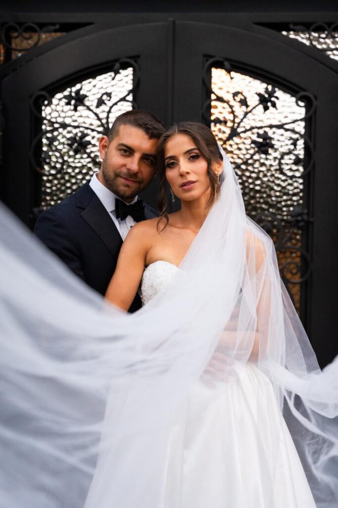 Bride and groom pose in front of ornate wrought-iron doors as her veil sweeps around them
