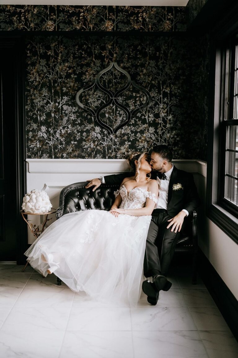 Bride and groom lounge on a tufted sofa in a room with dark damask wallpaper